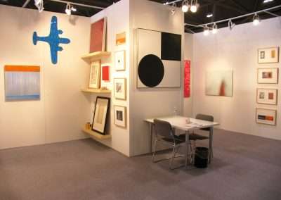 An art gallery room with white walls displaying various framed artworks, including abstract paintings and graphic designs. A small table with two chairs and a water bottle is set up in the middle. A blue wall-mounted sculpture shaped like an airplane is on the left.