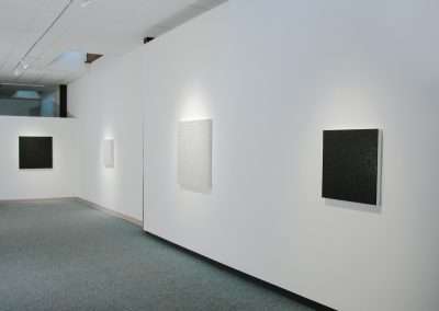 A minimalist art gallery featuring four square abstract paintings by Teo Gonzalez hung on a white wall. The paintings are monochromatic, varying in shades of black and white. The gallery space is well-lit with track lighting, providing a clean and modern atmosphere.