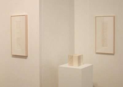 A minimalist art gallery features two framed monochromatic artworks on opposite walls and a small cube sculpture on a white pedestal in the center. The space is brightly lit with a soft, neutral-colored background.