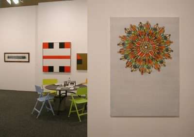 An art gallery displays colorful, geometric paintings. A round table with four differently colored chairs is set up in front of the artwork. A large, circular, multicolored piece is prominently hung on a white wall to the right.