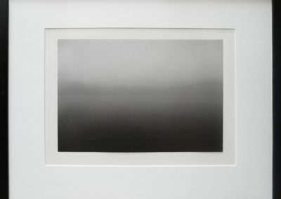 A framed artwork features a minimalist black and white gradient, transitioning smoothly from light gray at the top to darker shades at the bottom. The frame is black with a white matte border, offering a stark contrast to the muted tones of the image.