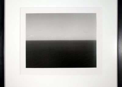 A black and white framed photograph depicting a calm, expansive sea under a cloudy sky. The horizon line is centered, dividing the ocean below and the sky above. The simplicity of the scene evokes a sense of tranquility and vastness.
