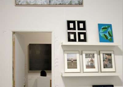 A gallery room with various artworks. Above a doorway, there is an abstract painting. To the right, a small shelf displaying framed photos, paintings, and a globe. Above the shelf are a grid of four black and white pictures and a colorful abstract painting.