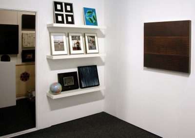 A small art gallery room with white walls. Three wall-mounted shelves display a variety of framed artworks and photographs. A single larger artwork is hung on the adjacent wall. The carpet is dark and there is an open doorway leading to another room.