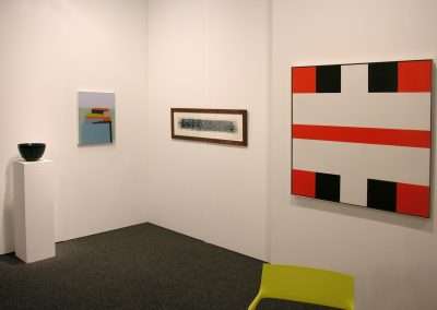 A gallery room displaying four artworks: a small modern painting with stacked shapes, a horizontal framed print, a large abstract canvas with red, black, and white geometric patterns, and a black bowl on a white pedestal. The room has neutral walls and carpeting.