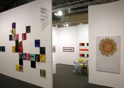 A gallery booth with various colorful abstract artworks displayed on white walls. The left wall features a collage of 3D mixed media pieces, while the right wall displays a large, circular, multi-colored painting. A small table with chairs is in the booth's corner.