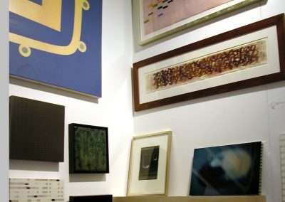 A gallery wall displaying various modern artworks, including framed paintings, a rectangular abstract piece, and a long, horizontal artwork with a looping pattern. Shelves below hold additional art pieces and books, creating a visually engaging display.