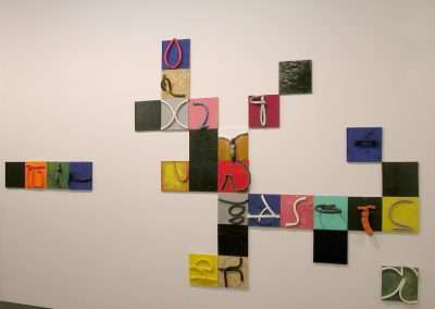 An art installation features a series of small, colorful square canvases arranged unevenly on a white wall. Each canvas displays a distinct, abstract pattern or shape in vibrant hues such as yellow, red, blue, green, and black.