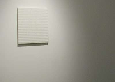 A minimalist art piece hangs on a plain, light-gray wall in a gallery. The artwork features a square canvas with subtle horizontal stripes and a monochromatic white color palette. The room is dimly lit, with a spotlight focused on the piece.