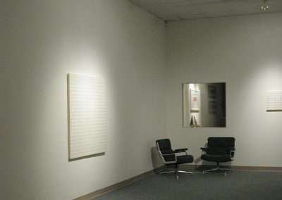 A minimalist art gallery room featuring two black chairs and a small table in one corner, and two abstract artworks on the walls. The room is softly lit with ceiling lights, highlighting the simplicity and clean lines of the artwork and furniture.