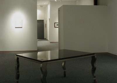 A minimalist art gallery features a black table with twisted metal legs on caster wheels as the central piece. Some framed artworks are displayed on the white walls. The setting is simple, with track lighting illuminating select areas.