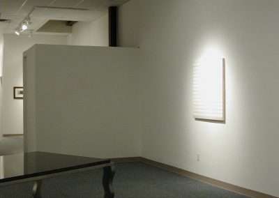 An art gallery displaying a single minimalist piece on a white wall. The artwork features white horizontal lines on a white canvas. Nearby, there is a glass-topped table with curved metal legs and wheels. The space is lit by overhead track lighting.