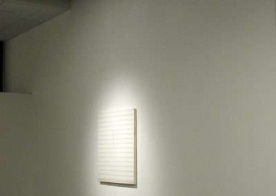 A large, nearly empty room with a single illuminated piece of art on one of the walls. The artwork is minimalistic, consisting of faint horizontal and vertical lines, and is highlighted by a focused spotlight. The rest of the room and walls are plain and unadorned.