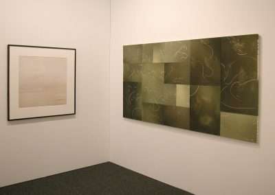 A minimalist art gallery corner featuring two abstract pieces: one framed monochrome artwork to the left and a larger, multi-panel greenish-gray piece with subtle abstract designs on the right. Both hang on plain white walls.
