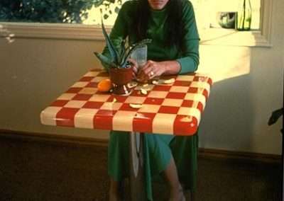A person wearing a green dress sits barefoot at a small table with a red and white checkered tablecloth. The table holds a potted plant, an orange, and a drink. They are inside near a window with greenery and a vase with orange flowers visible in the background.