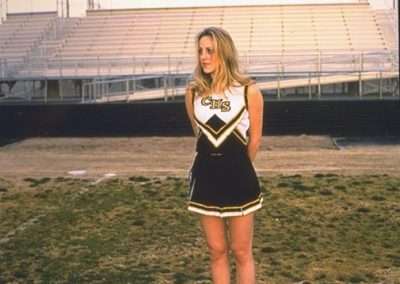 A cheerleader in a black and yellow cheerleading uniform stands on a grassy field in front of empty stadium bleachers. She has her hands behind her back and is looking slightly to her left. The sky is cloudy.