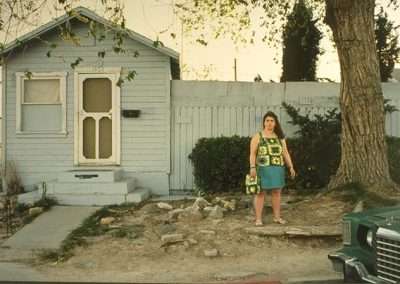 A woman stands in front of a small, weathered house with a screen door and peeling paint. She wears a green and yellow floral top, a blue skirt, and holds a matching bag. A large tree is beside her and a green car is partially visible in the foreground.