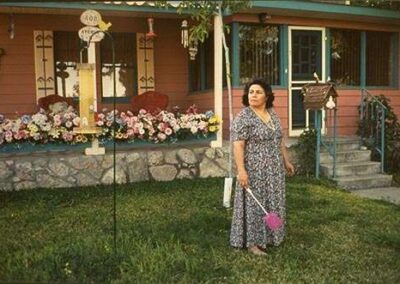 A woman in a patterned dress stands holding a pink duster in the yard of a house with a large porch. The house is decorated with flowers and has a stone planter in front. Various garden ornaments are also visible.