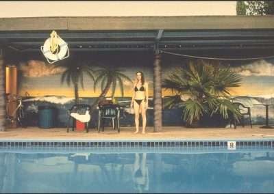 A woman stands by the edge of a swimming pool in a bikini. Behind her is a mural depicting a beach scene with palm trees and a sunset on the wall of a poolside building. Surrounding her are various items including bikes, a life preserver, and a palm plant.