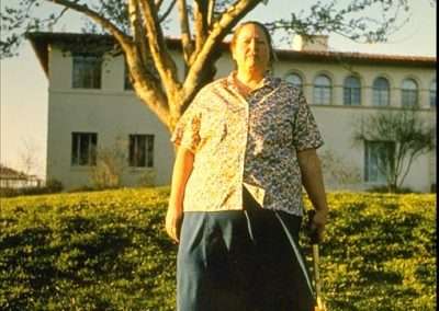 A person stands on a grassy lawn holding a cane with one arm extended downward. They are wearing a patterned short-sleeve shirt, blue skirt, and white shoes. A large tree and a light-colored building are in the background under a clear sky.
