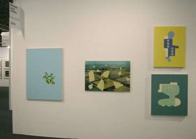 An art gallery wall displays four paintings. From left to right: an abstract design with green shapes on a light blue background, an aerial landscape, and two paintings with abstract forms on light yellow and green backgrounds. A nameplate for Richard Levy Gallery hangs above.