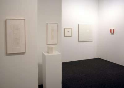 A modern art gallery features minimalistic artwork. On the left wall, there are two framed sketches. A small white sculpture sits on a pedestal. The back wall displays three pieces: a small framed drawing, a gridded painting, and a piece with vertical stripes.
