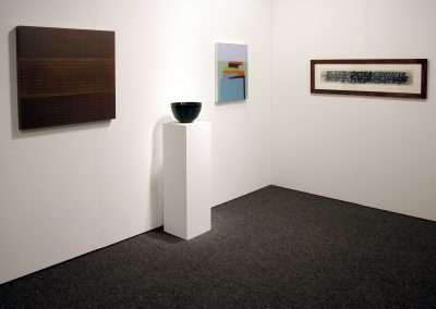 An art gallery corner displaying a wooden panel artwork, a multicolored geometric painting, a ceramic bowl on a white pedestal, and a long framed monochrome piece. The floor is carpeted and the walls are white.