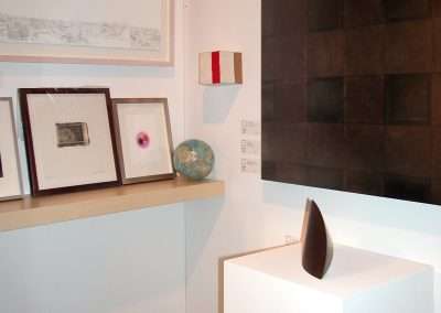 An art gallery display features various framed artworks and a colorful globe on a shelf, with a geometric black and brown wall piece to the right and a minimalistic sculpture on a white pedestal in the foreground.