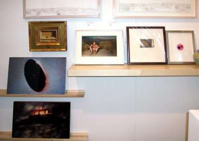 A gallery wall with various framed artworks displayed on shelves and hooks. The pieces include photographs, a small abstract painting, and a few other art prints. One photo depicts a person sitting outside, while another features a dark circular shape.
