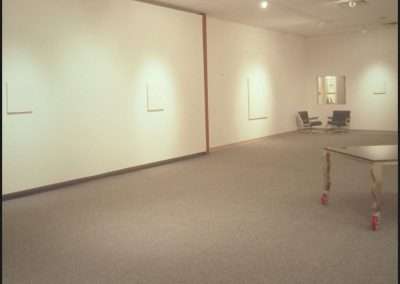 A spacious art gallery room with carpeted flooring. The walls are mostly bare, except for a few small, unidentifiable objects or artworks. There are also a few chairs and a table with wheels in the middle of the room. The area is well-lit by ceiling lights.