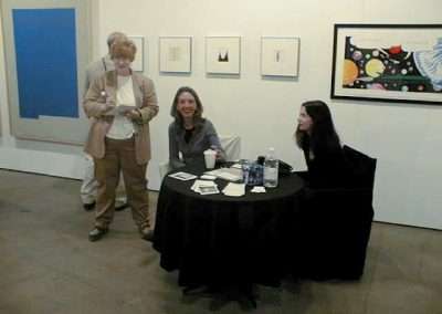 Three people are gathered in an art gallery. Two of them are seated at a round table with black covering, which has beverages and papers on it. The third person stands nearby holding something. The gallery walls display various framed artworks.