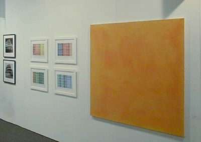 An art gallery displays several pieces on a white wall. On the left, there are two vertical photo frames followed by two sets of four smaller framed images with colorful grid patterns. To the right, a large square painting in varying shades of orange is prominently featured.
