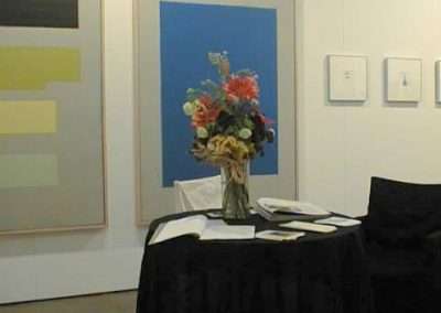 A small round table with a black cloth stands in front of a flower arrangement in a vase. The table is surrounded by books and papers. Behind it, various abstract paintings are displayed on the walls of a gallery. A dark chair is partially visible to the right.
