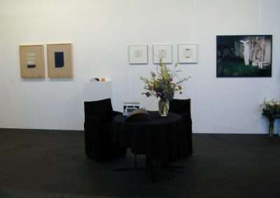 An art gallery exhibit with four framed artworks on the wall. In the center, a round table covered with a black cloth holds a large bouquet of flowers. The gallery has minimal decoration, and a small flower arrangement is on the floor in the corner.