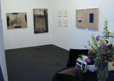 A minimalistic art gallery room featuring four abstract paintings on white walls. The paintings are in neutral tones of black, beige, and gray. A table with purple and yellow flower arrangements, along with some papers, is visible in the foreground.