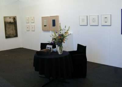 A minimalist exhibition space with framed artwork on white walls. A small round table draped with a black cloth holds a bouquet of flowers and a small device, flanked by two black chairs. The room has a clean, simple design with a quiet, contemplative atmosphere.