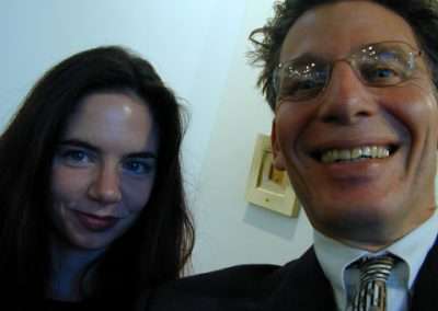 A close-up photo of a woman with long dark hair and a subtle smile standing next to a man with glasses, smiling widely. The man is wearing a suit and tie. They appear to be indoors with a light-colored wall in the background.