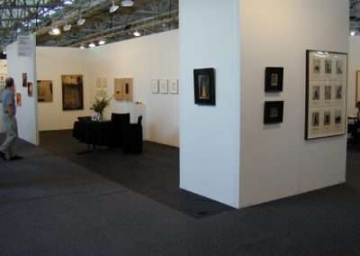 An art gallery with white walls displays framed artwork, including photographs and paintings. A narrow table with a black tablecloth holds a floral arrangement. A few visitors can be seen walking around, viewing the exhibits under the hanging ceiling lights.