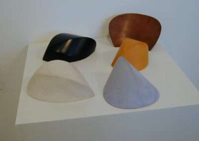 A photo of five conical sculptures displayed on a white pedestal. Each cone is a different color: black, orange-brown, yellow, and two shades of white or light gray. The sculptures appear to be made of smooth, solid material.