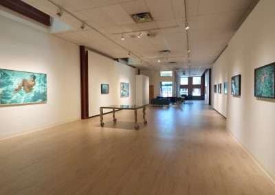 A spacious art gallery with a light wooden floor and white walls displays several framed paintings depicting water scenes. The room is illuminated by ceiling lights. In the center, a modern glass table on wheels is positioned near some seating.