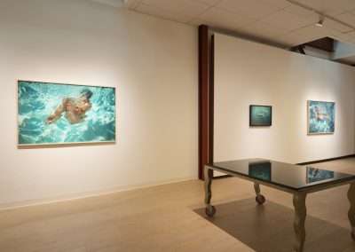 A modern art gallery displays framed artwork on white walls, featuring images of swimmers in water. A large dark-colored table with uniquely shaped legs stands in the center of the room on a light wood floor. Track lighting illuminates the space.