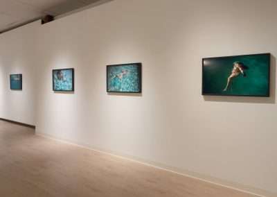 An art gallery wall displays four framed photographs of people swimming in clear blue water. Each image captures different underwater scenes and poses. The gallery room has a light wooden floor and plain, off-white walls. Soft lighting highlights the photographs.