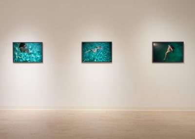 A minimalistic art gallery wall displaying three framed photographs, each depicting individuals swimming underwater in clear blue water. The gallery has wooden flooring and soft lighting focused on the artwork.