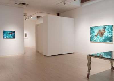 A modern art gallery features several pieces. On the left wall is a small painting of a swimmer. In the center is a larger framed artwork depicting a person swimming underwater in blue-green water. A reflective table sits on the right side of the room.