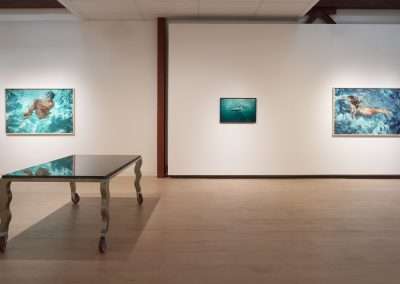 An art gallery displaying three paintings of figures swimming underwater. The paintings are mounted on a white wall. In the foreground, there is a reflective metal table on wheels with wavy legs. The floor is light wood, contributing to the minimalist setting.