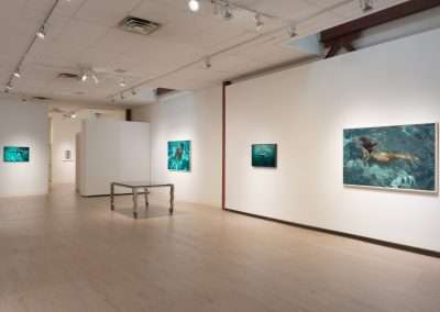 A modern art gallery with white walls displays several framed paintings of underwater scenes. A minimalist metal table stands in the center of the room. Recessed ceiling lights illuminate the artwork, and a doorway at the end leads to another room.