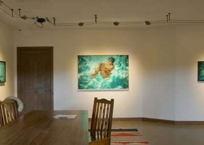 An art gallery room at Bishop's Lodge features three paintings of swimmers in a pool by Eric Tillinghast. The largest painting is centered on the main wall, with two smaller ones flanking it. A wooden dining table and chairs occupy the foreground, while a potted plant rests in the left corner.