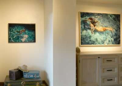 A room at Bishop's Lodge features two paintings of swimmers on the wall, separated by a doorway. Below Eric Tillinghast’s painting on the left are two suitcases and a hat, while beneath Manjari Sharma’s artwork on the right stands a wooden dresser with a small potted plant on top.
