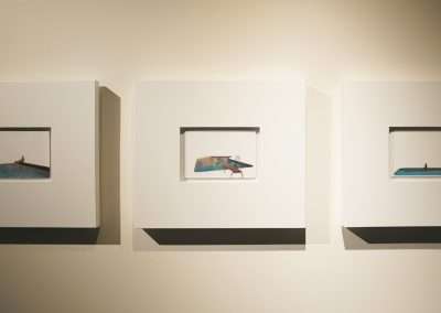 A photograph of an art display at Bishop's Lodge shows three framed images hung on a wall. Each frame contains a small, minimalist painting by Eric Tillinghast, predominantly featuring shades of blue and depicting swimming pools with simple, abstract human figures. The frames are evenly spaced.
