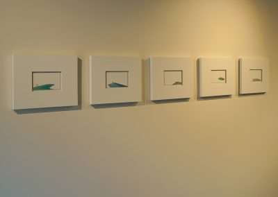 A wall at Bishop's Lodge features five small, square, white-framed artworks arranged in a horizontal line. Each frame contains a minimalist image of a colored paper plane placed on a blank white background. The paper planes are various shades of blue, green, and white.
